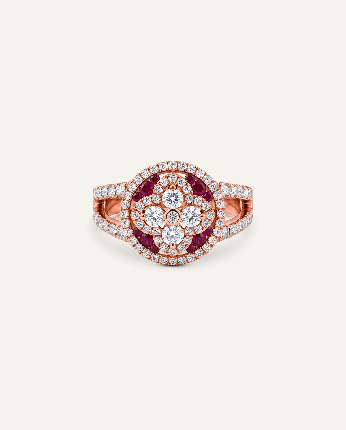 A rose gold ring featuring a circular floral cluster design with dark red round-cut stones and surrounding clear pavÃ© stones, set against a solid off-white background.