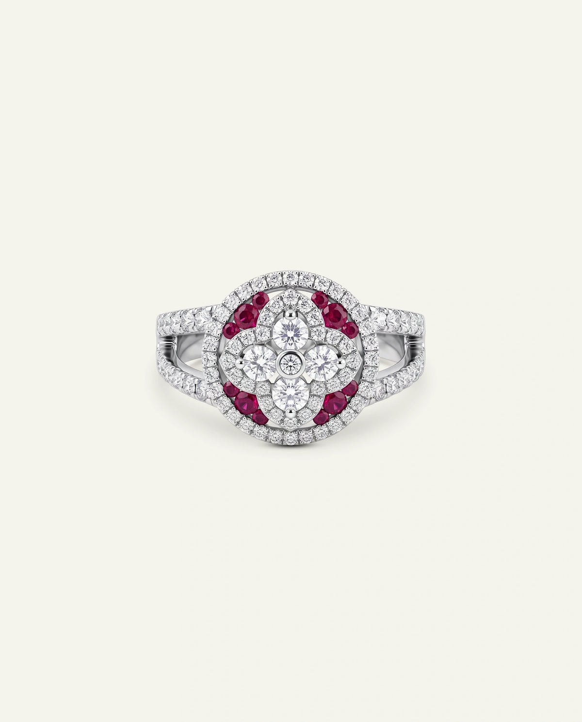 A silver-toned oval ring featuring a floral cluster center with a single round clear stone surrounded by smaller clear stones, bordered by a halo of deep red gemstones, with a pavÃ©-set band of clear stones, shown on a light cream background.