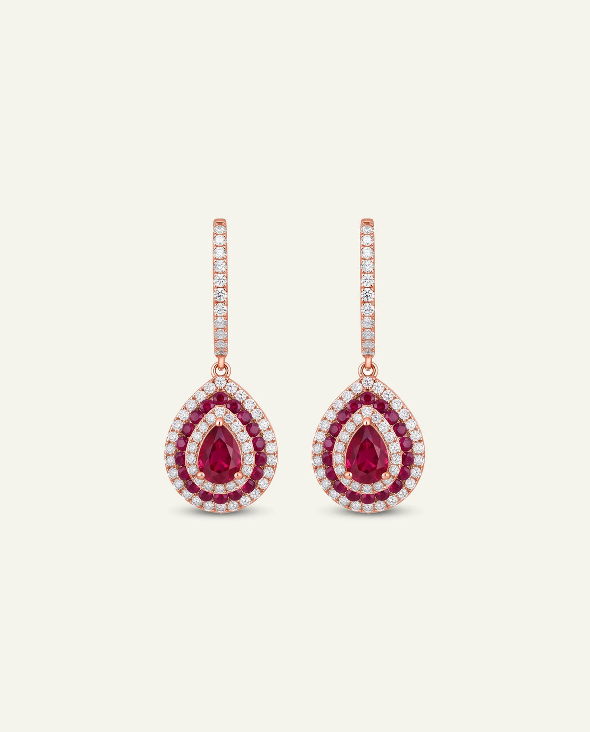 A pair of rose gold drop earrings featuring teardrop-shaped ruby gemstones surrounded by pavé-set clear stones, shown against a light cream background.