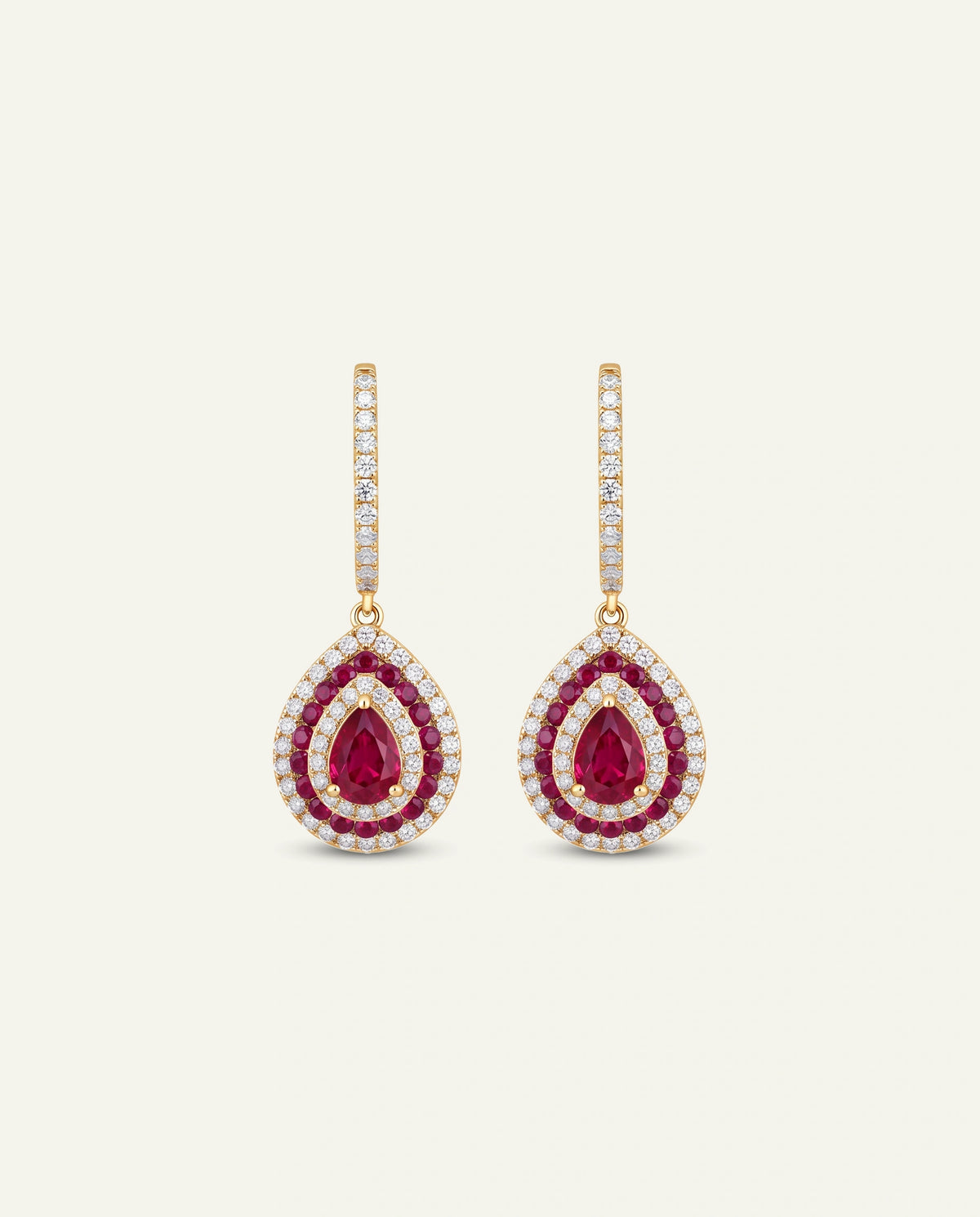 A pair of gold teardrop drop earrings featuring a central round ruby gemstone surrounded by a halo of small clear stones, with a vertical bar of clear stones above, set against a light cream background.