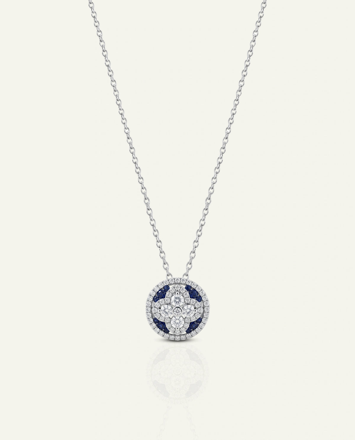 A delicate silver chain necklace featuring a circular pendant with a silver-toned cross design, accented by deep blue stones and surrounded by small clear stones, set against a light cream background.