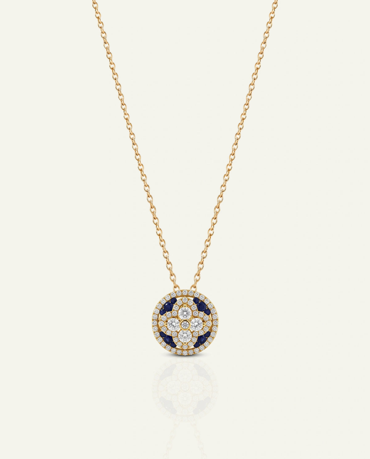 A yellow gold chain necklace featuring a circular pendant with a center four-point cross motif set with clear diamonds and deep blue sapphire accents, enclosed by a diamond halo, shown on a light cream background.