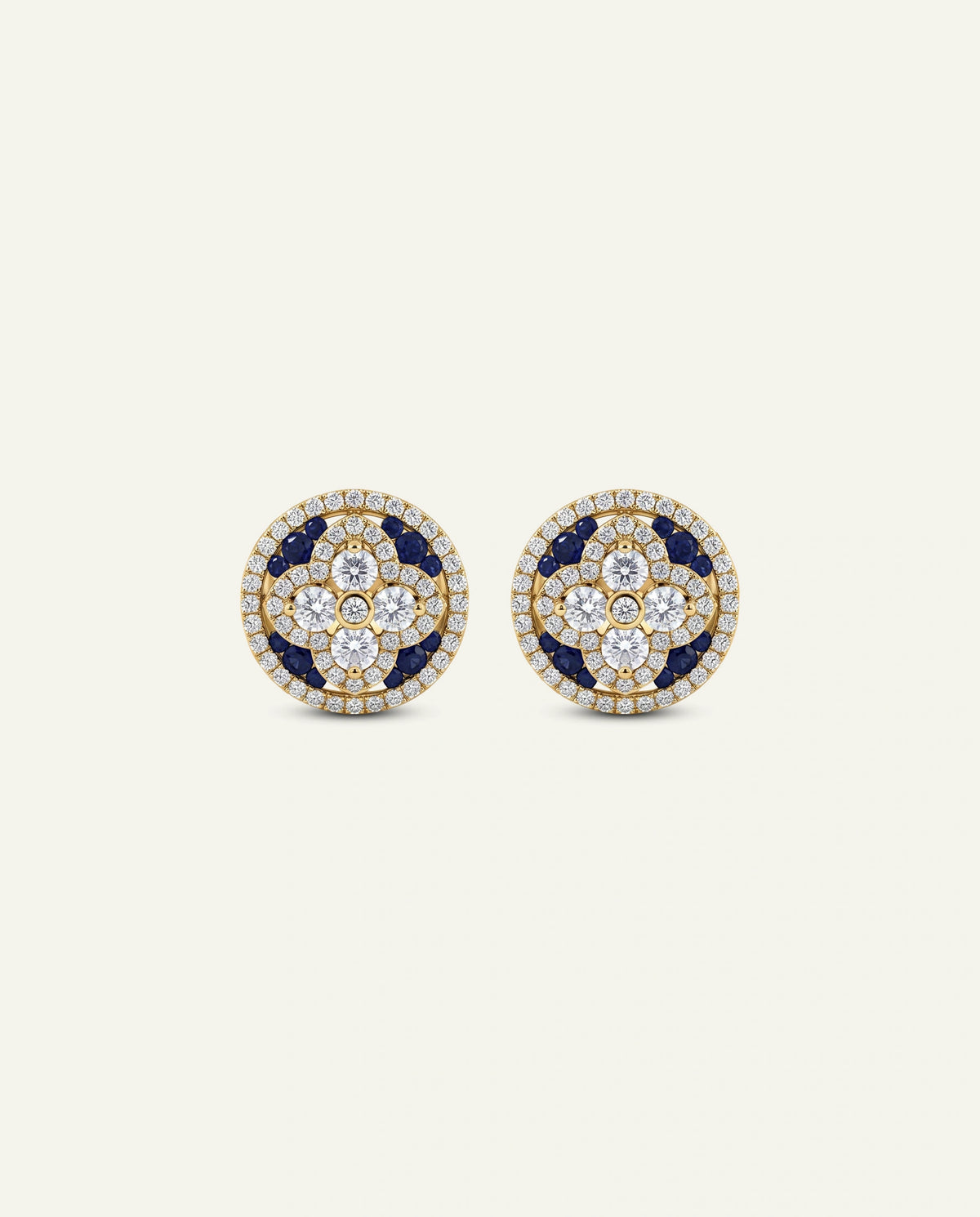 A pair of round gold stud earrings featuring a floral pattern of gold, deep blue, and clear stones, set against a solid off-white background.