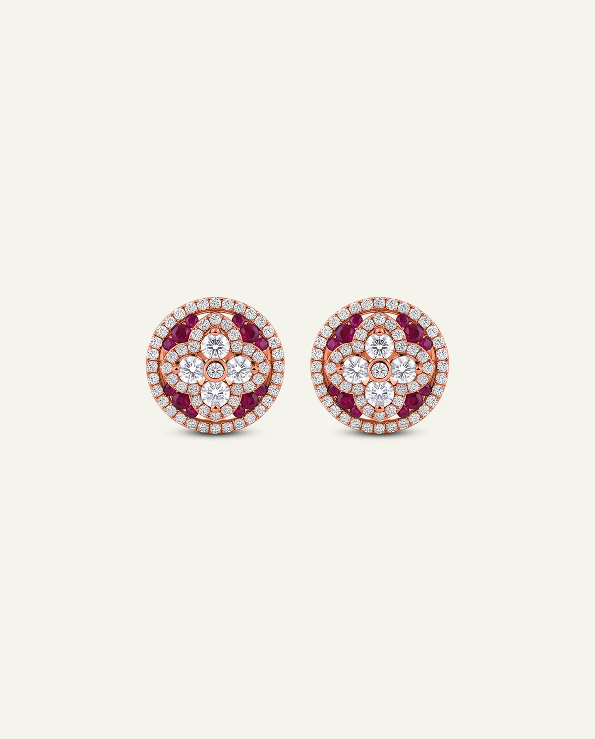 A pair of round rose gold stud earrings decorated with concentric circles of clear pavÃ© diamonds and deep red gemstones, set against a solid off-white background.