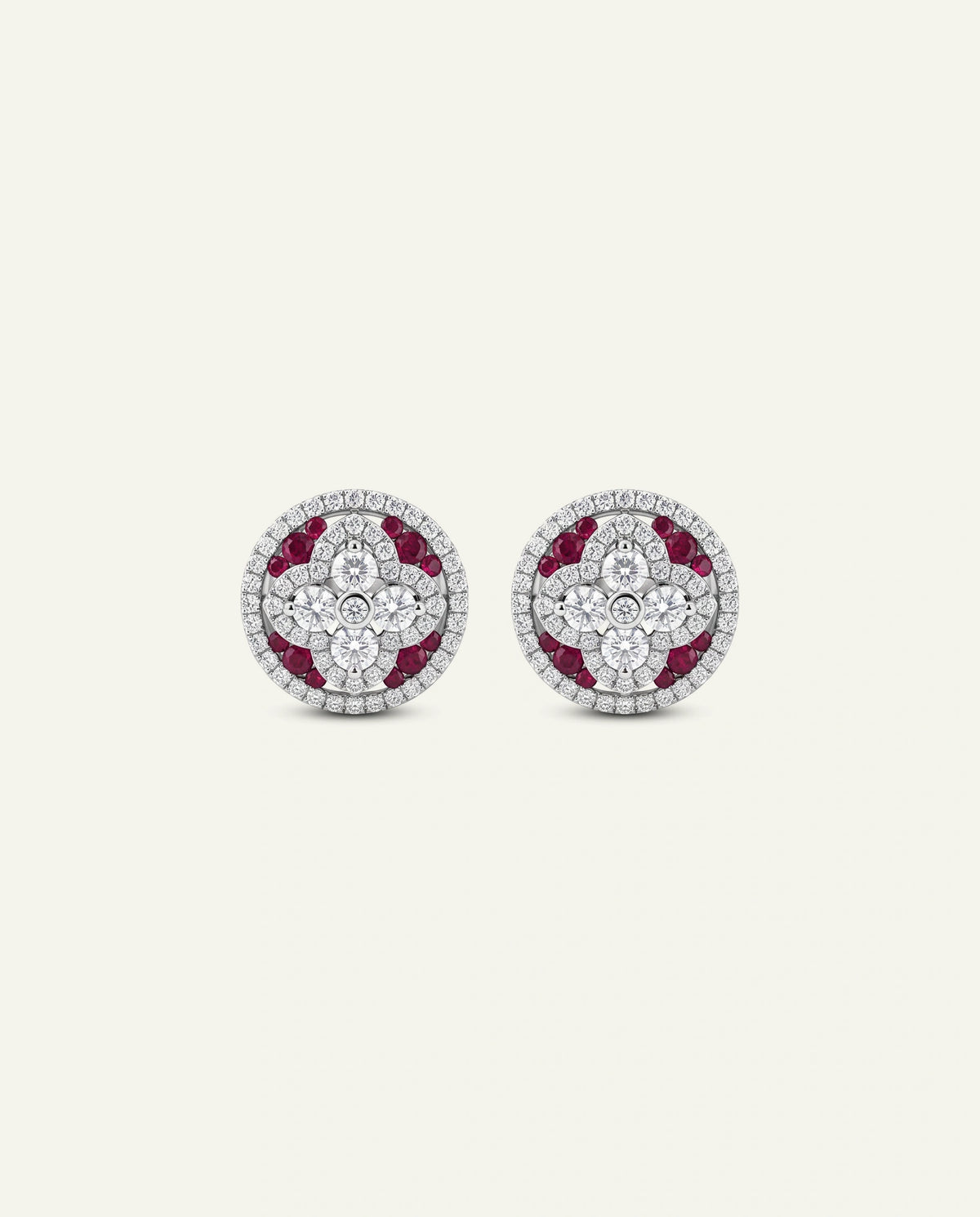 A pair of white gold round stud earrings featuring a center diamond cluster in a four-petal floral design, bordered by vibrant red ruby accents and an outer halo of pavÃ©-set diamonds, shown on a light cream background.
