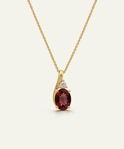 A gold pendant necklace featuring an oval deep red garnet stone and a small clear accent stone, displayed on a clean off-white background.