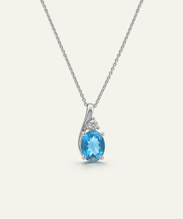 A silver pendant necklace featuring an oval-cut light blue topaz stone in a prong setting, accented by small round clear stones above it, suspended from a delicate silver chain against a light cream background.