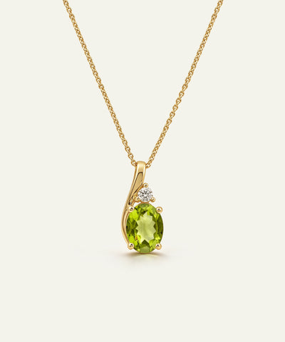 A gold pendant necklace featuring an oval-cut green peridot stone and a small clear accent stone, shown on a delicate gold chain against a light cream background.