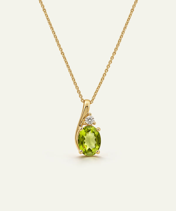 A gold pendant necklace featuring an oval-cut green peridot stone and a small clear accent stone, shown on a delicate gold chain against a light cream background.