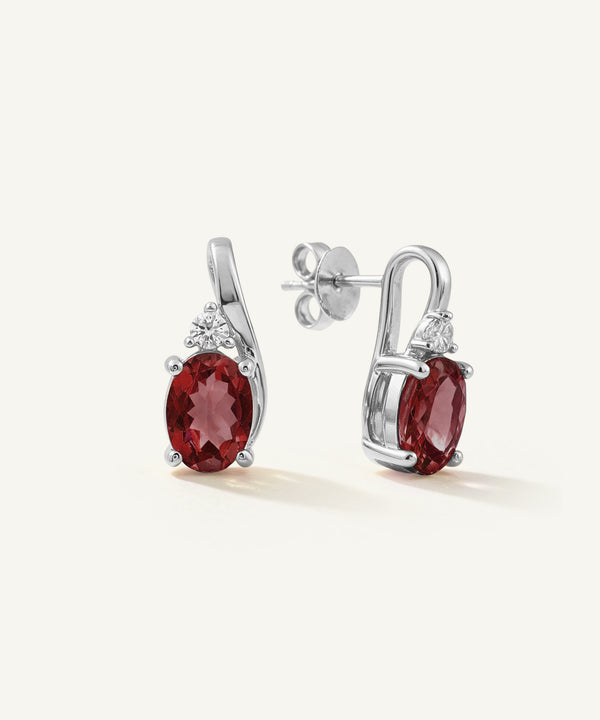 A pair of silver-toned drop earrings featuring oval deep red gemstones and small clear accent stones, displayed on a clean off-white background.