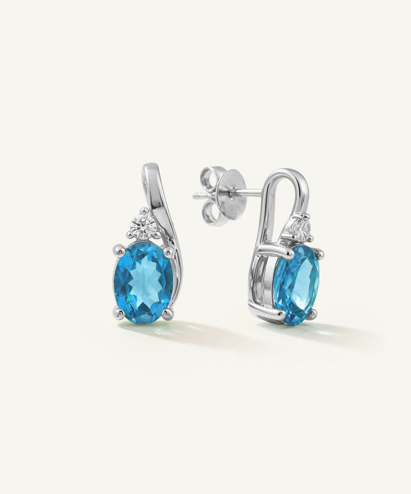 A pair of silver drop earrings featuring oval-cut light blue gemstones and small clear accent stones, shown on a clean off-white background.
