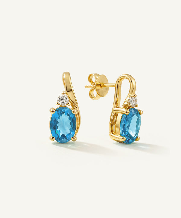 A pair of gold drop earrings featuring oval-cut blue topaz stones and small clear accent stones, displayed on a clean off-white background.