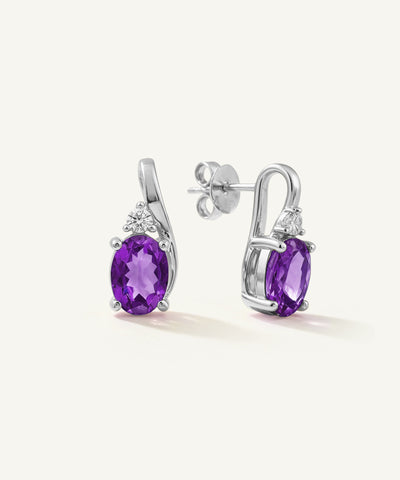 A pair of silver drop earrings featuring oval purple amethyst gemstones and small clear accent stones, shown on a clean off-white background. - alternate view