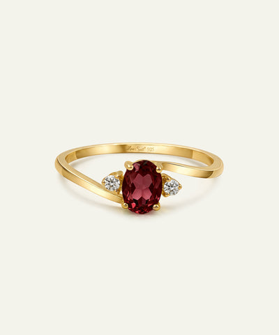 A yellow gold ring featuring an oval-cut deep red ruby center stone, flanked by two small round-cut clear diamonds on a light cream background.