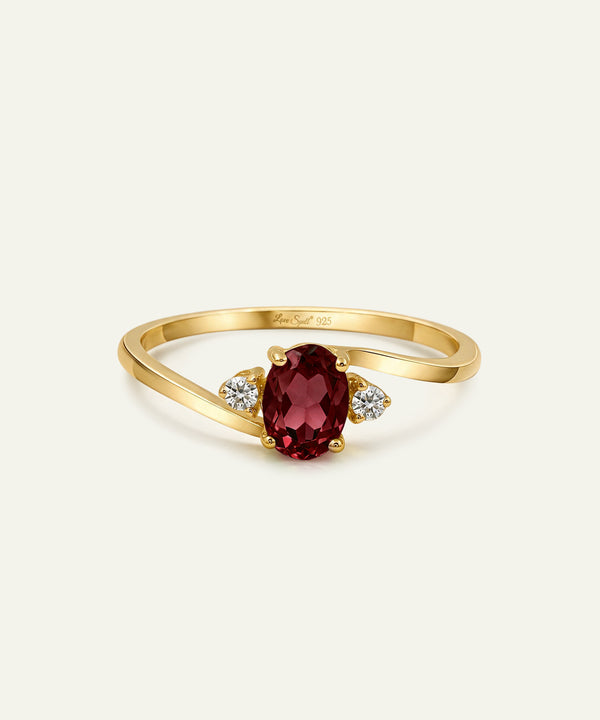 A yellow gold ring featuring an oval-cut deep red ruby center stone, flanked by two small round-cut clear diamonds on a light cream background.