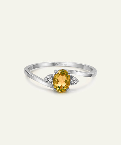 A silver ring featuring an oval-cut yellow citrine center stone, flanked by two small round-cut diamonds on a light cream background. - alternate view