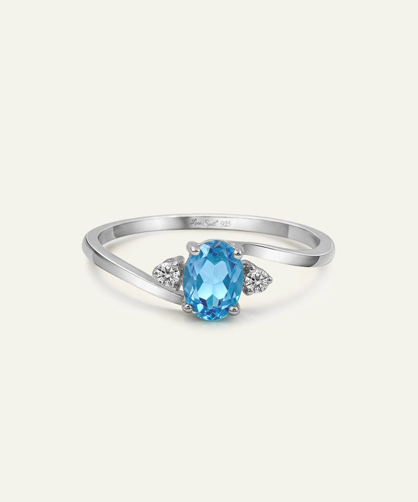 A silver ring featuring an oval-cut light blue gemstone in the center, flanked by two small round clear stones on a cream background.