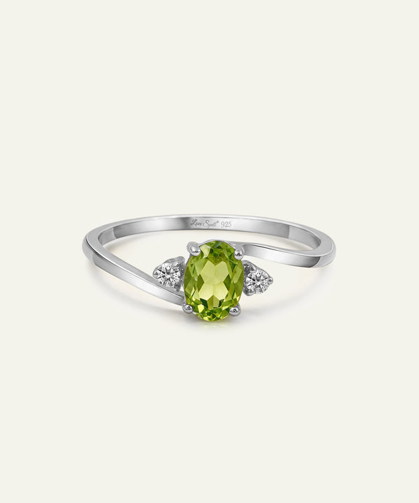 A silver ring featuring an oval-cut green peridot center stone and two small round-cut clear accent stones on either side of the band, shown on a light cream background.