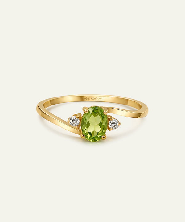 A yellow gold ring featuring an oval-cut green peridot center stone and two small round-cut clear accent stones on either side, set against a plain off-white background.