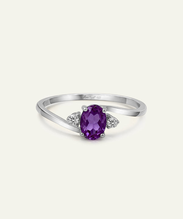 A silver ring featuring an oval-cut purple amethyst center stone, accented by two small round-cut clear stones on either side of the band, shown on a light cream background.