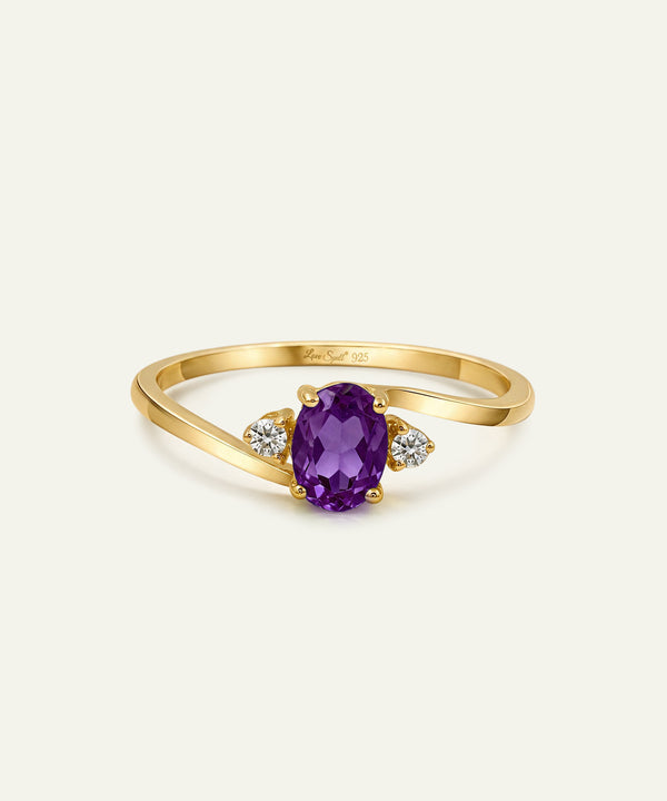 A yellow gold ring featuring an oval-cut purple amethyst center stone, flanked by two small round-cut clear diamonds on a light cream background.