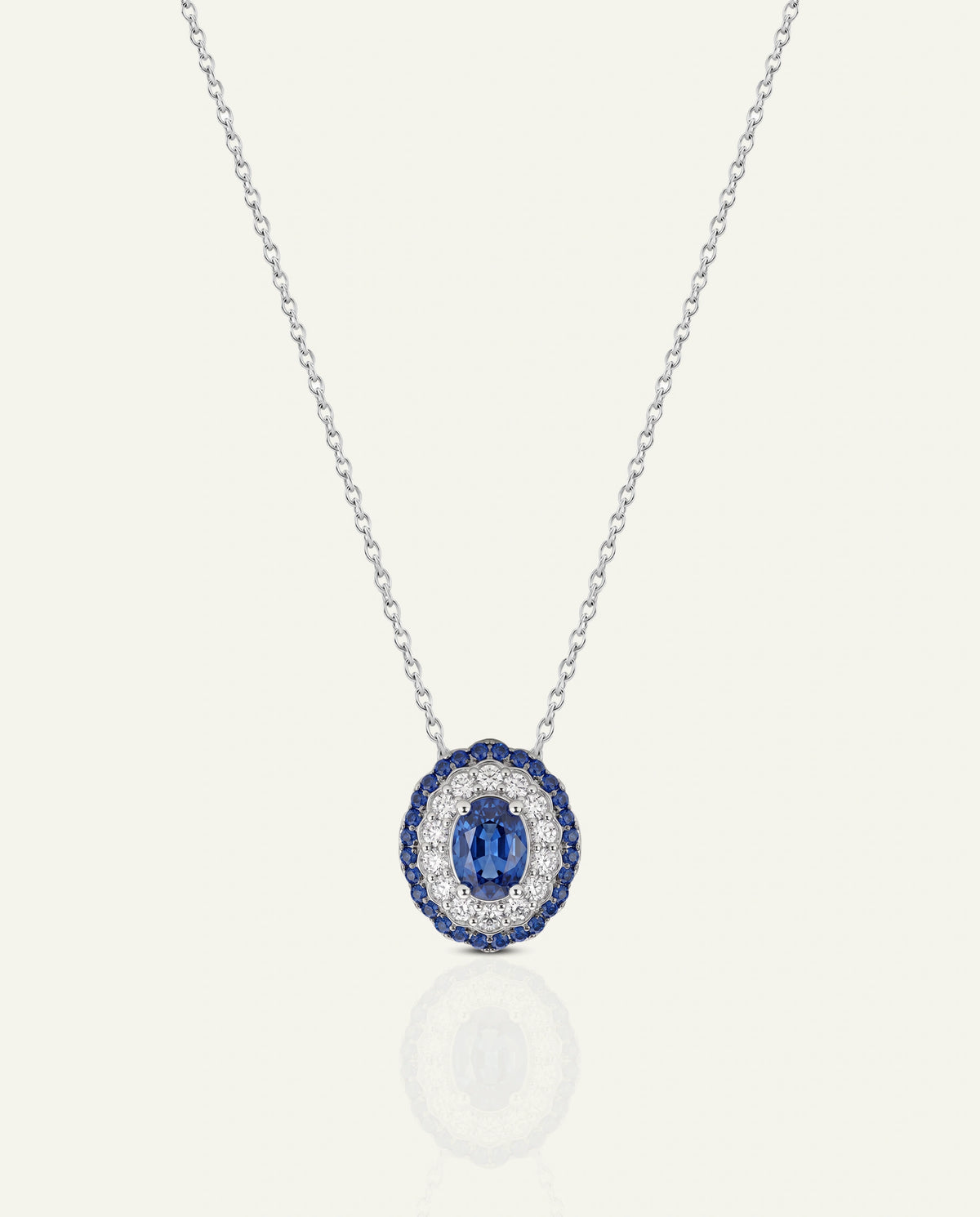 A silver-tone oval pendant necklace featuring a central deep blue oval gemstone surrounded by a halo of small clear stones, set against a light cream background with a soft reflection below.