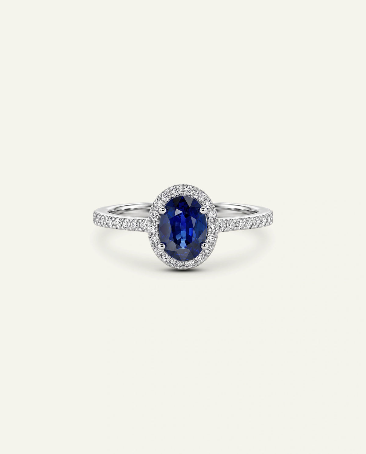 An oval-cut blue sapphire ring with a halo setting and a silver-colored band paved with small clear stones, centered on a light cream background.