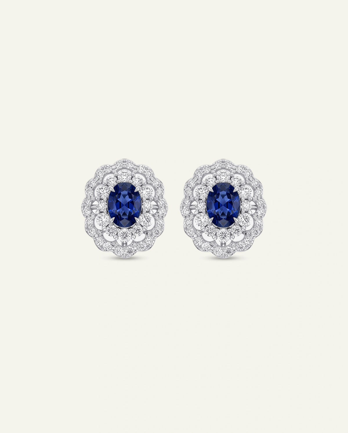 A pair of oval cluster stud earrings featuring deep blue oval-cut center stones surrounded by a halo of small clear stones in a silver-toned setting, shown on a plain light beige background.