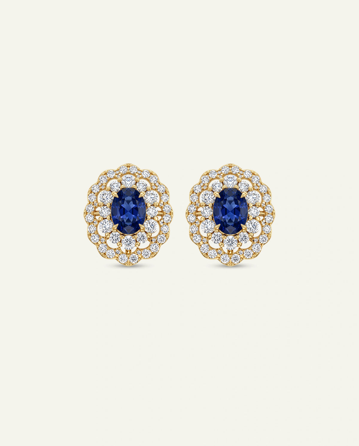 A pair of yellow gold stud earrings featuring central oval-cut blue sapphires framed by an intricate, two-tier scalloped halo of small round clear pavé diamonds, set against an off-white background.