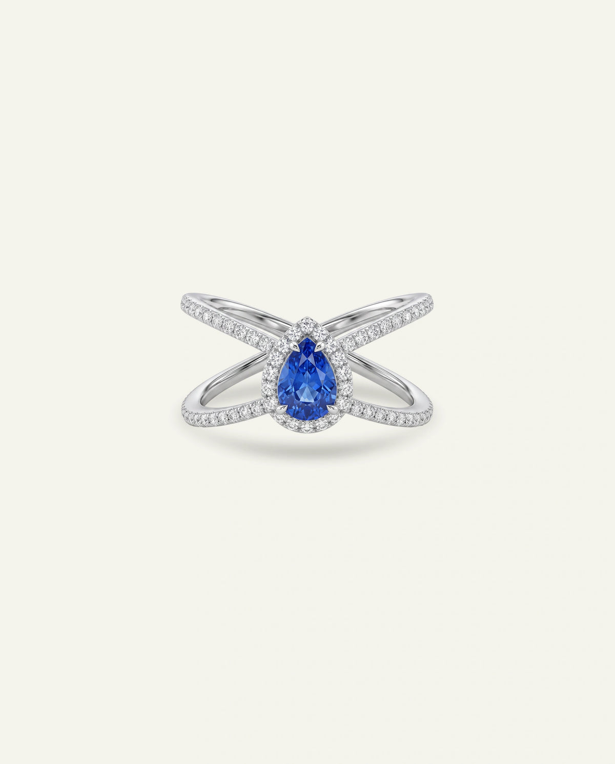 A silver-toned ring featuring a pear-cut blue sapphire gemstone in a halo setting, with crossed shanks paved with small clear stones, shown on a light cream background.