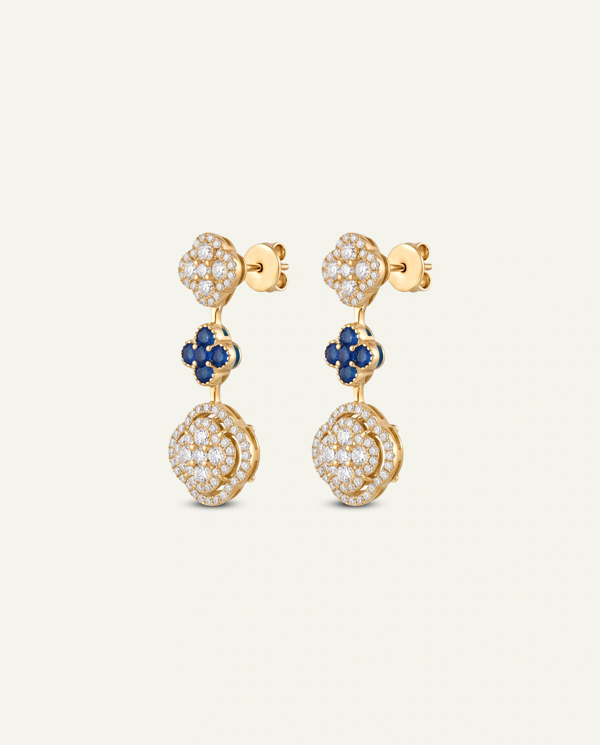 A pair of gold drop earrings featuring pavé-set clear stones, a small blue floral accent, and a larger round pavé stone at the bottom, shown on a light cream background.