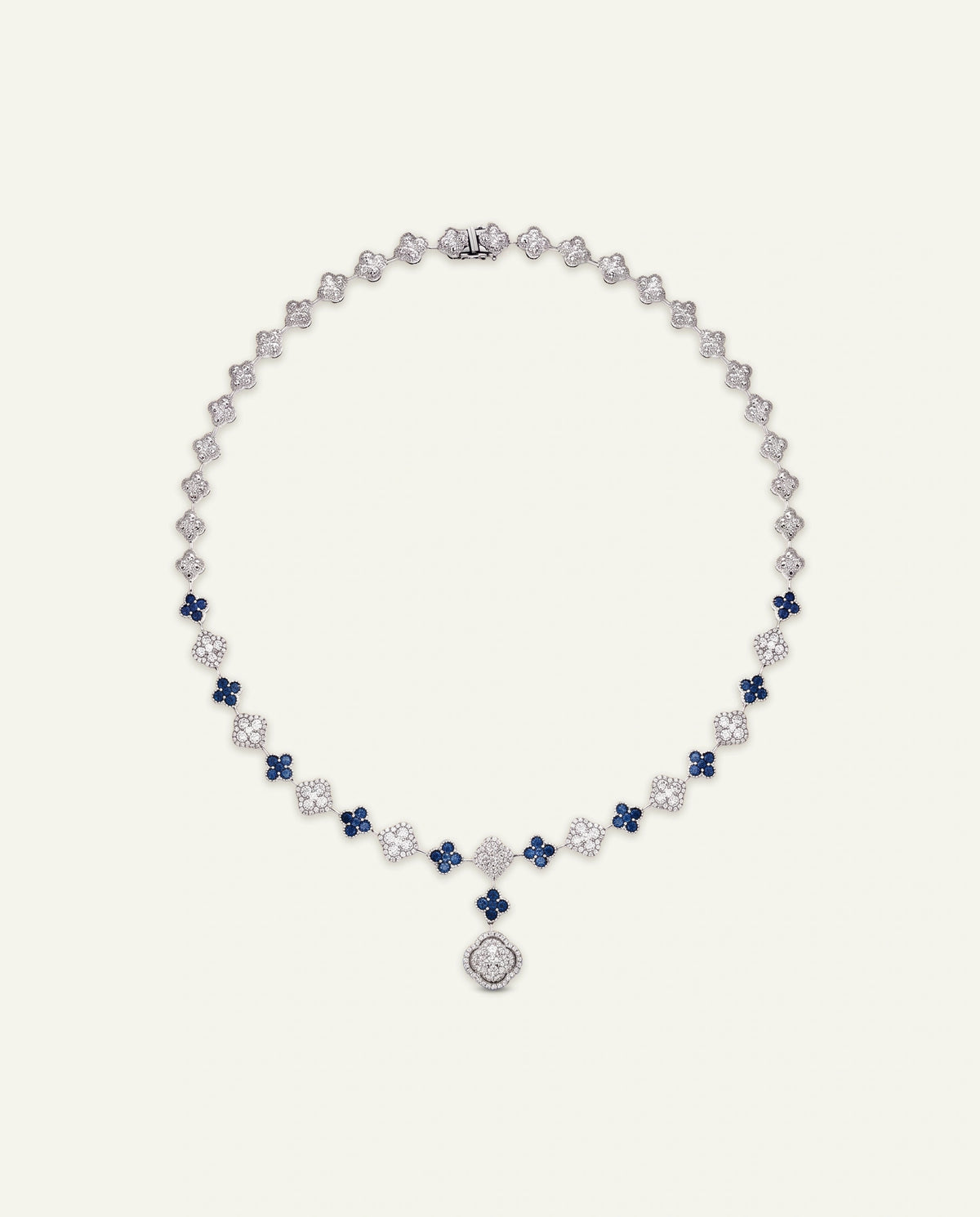 A luxury diamond and sapphire necklace with a floral clover motif and a central diamond medallion pendant, displayed on a light cream background.
