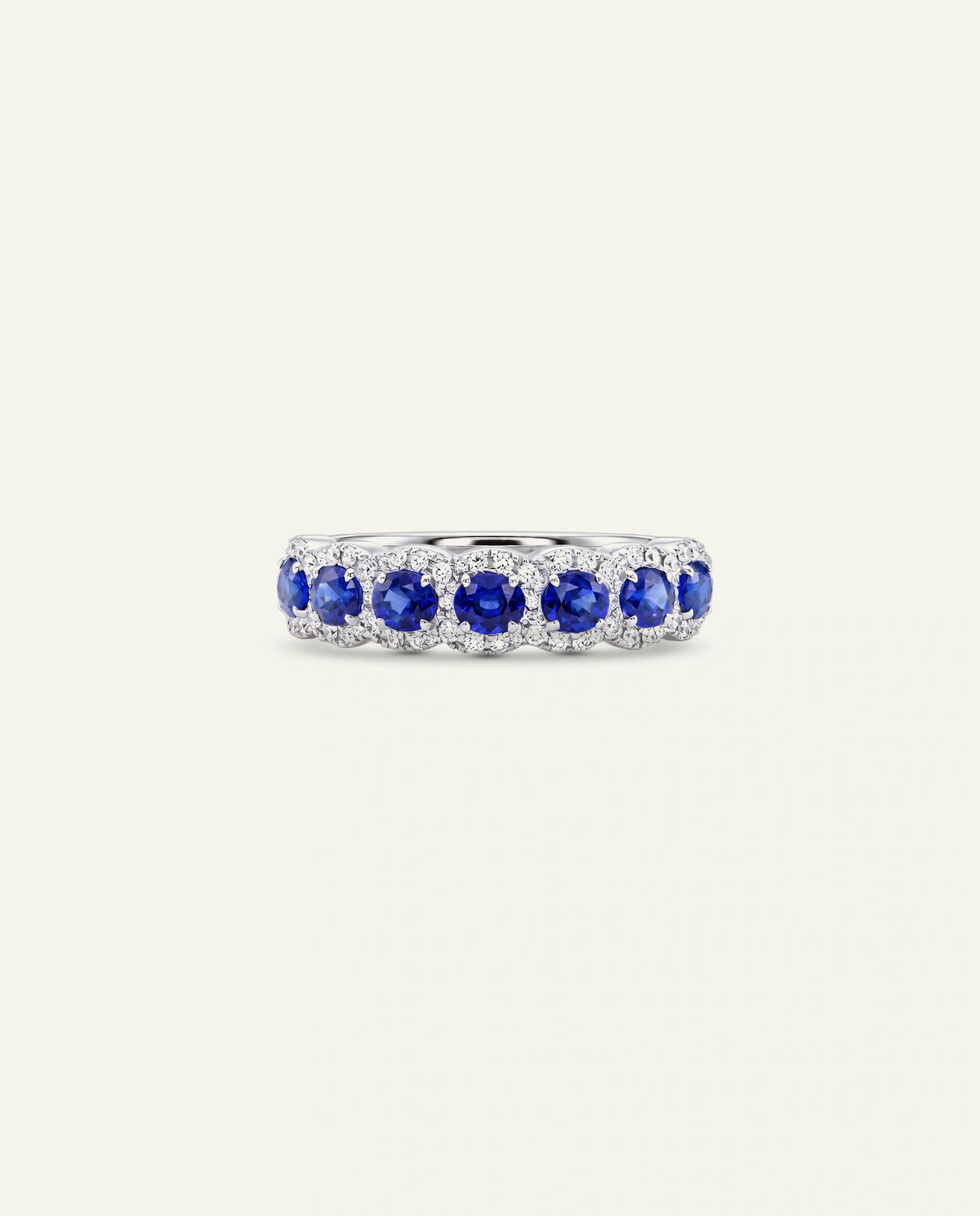 A silver band ring featuring a row of round-cut blue sapphires and small clear accent stones, set against a solid off-white background.