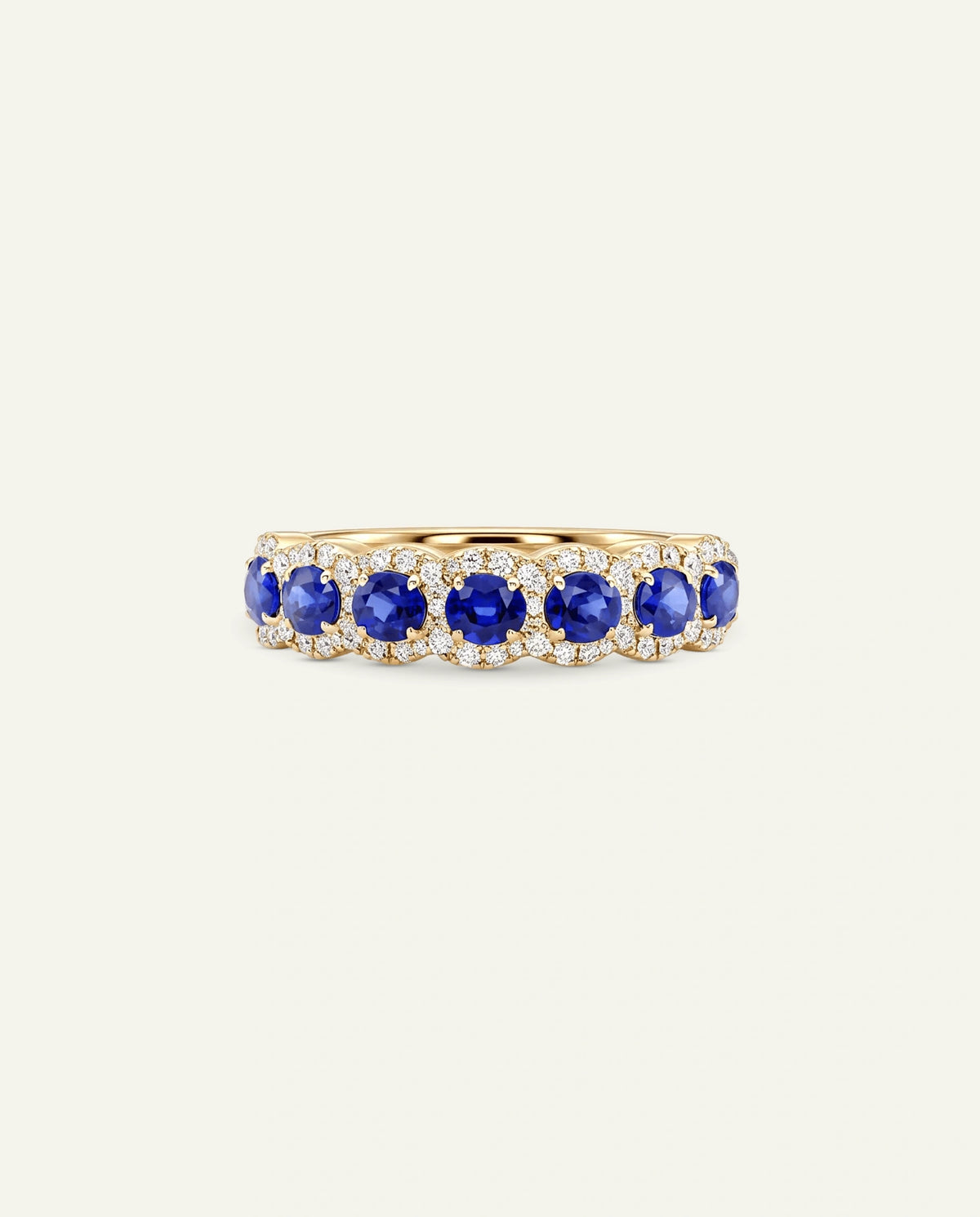 A gold eternity ring featuring five round-cut deep blue sapphires and small clear accent stones in a halo setting, centered on a light cream background.