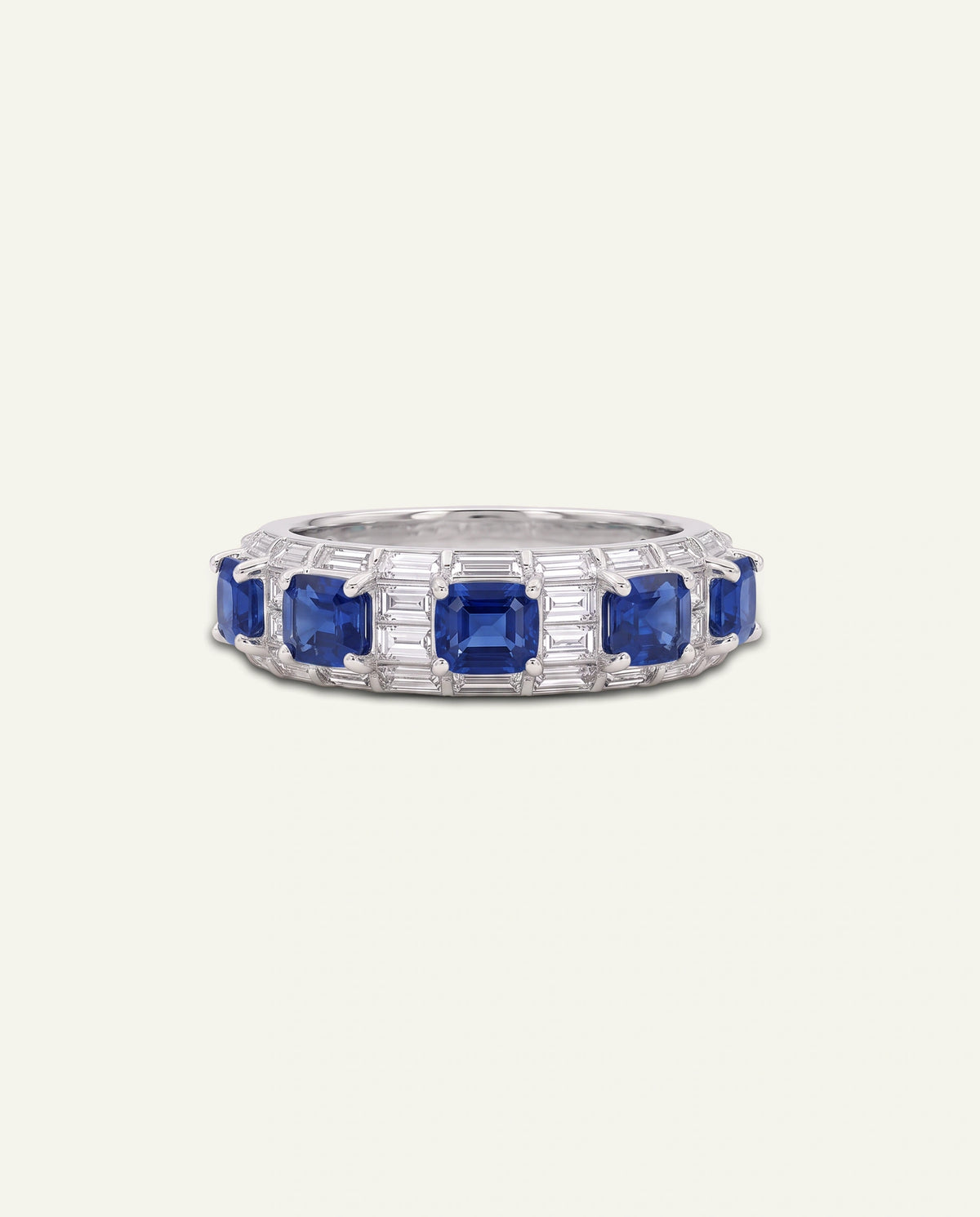 A silver band ring featuring a row of square-cut blue sapphires and baguette-cut clear stones, presented on a light cream background.