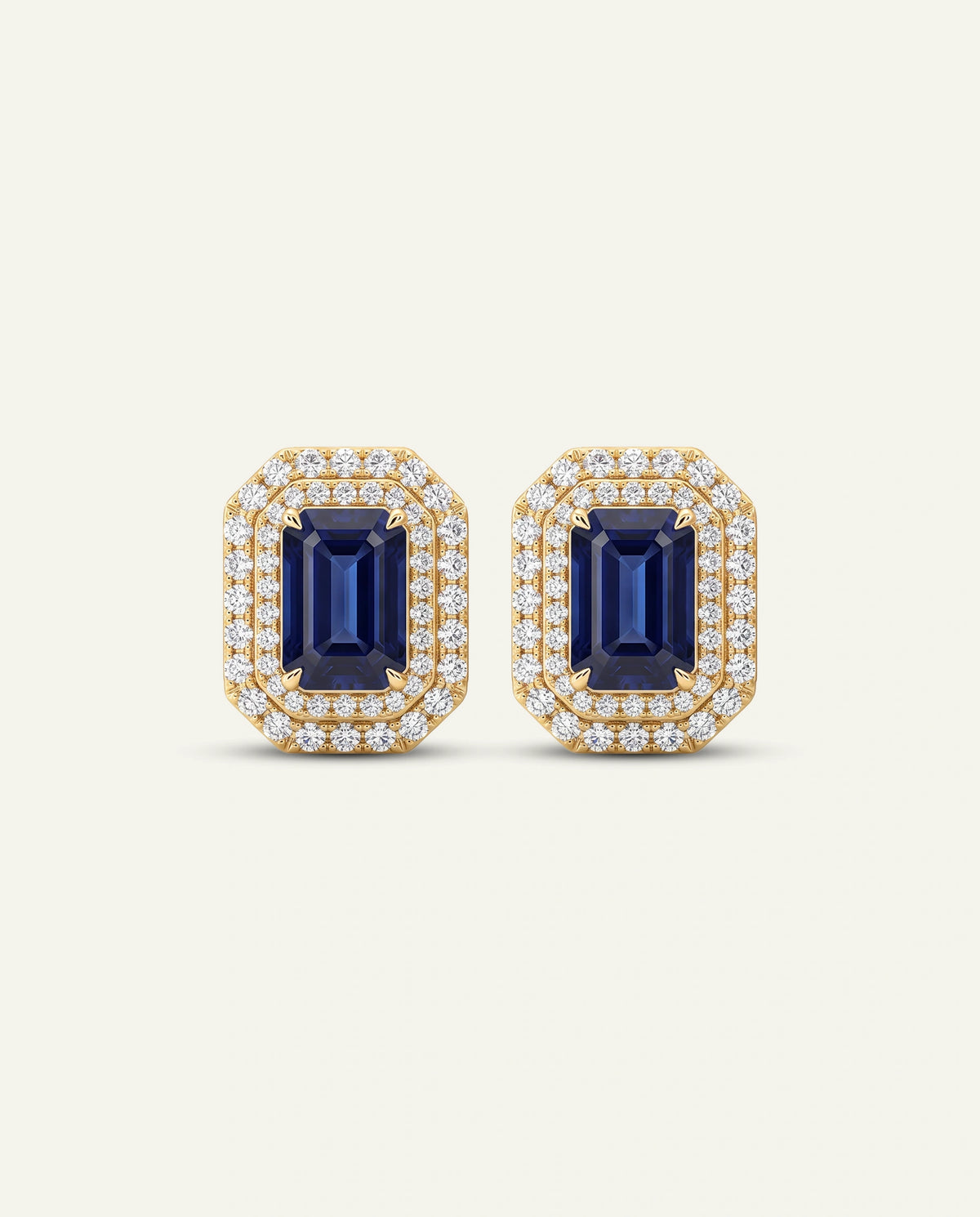A pair of yellow gold stud earrings featuring rectangular emerald-cut blue sapphires surrounded by double rows of small clear stones, set against a solid off-white background.