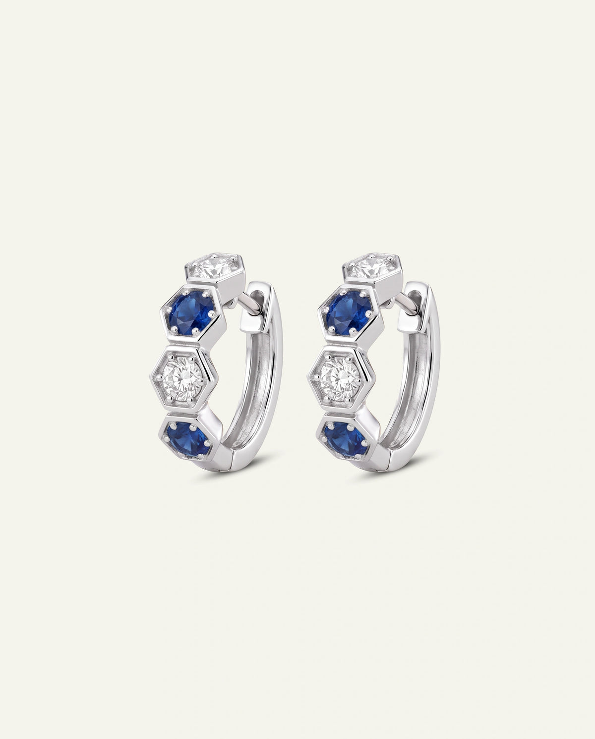 A pair of silver huggie hoop earrings featuring a honeycomb pattern with blue sapphires and clear cubic zirconia stones, set against a light cream background.