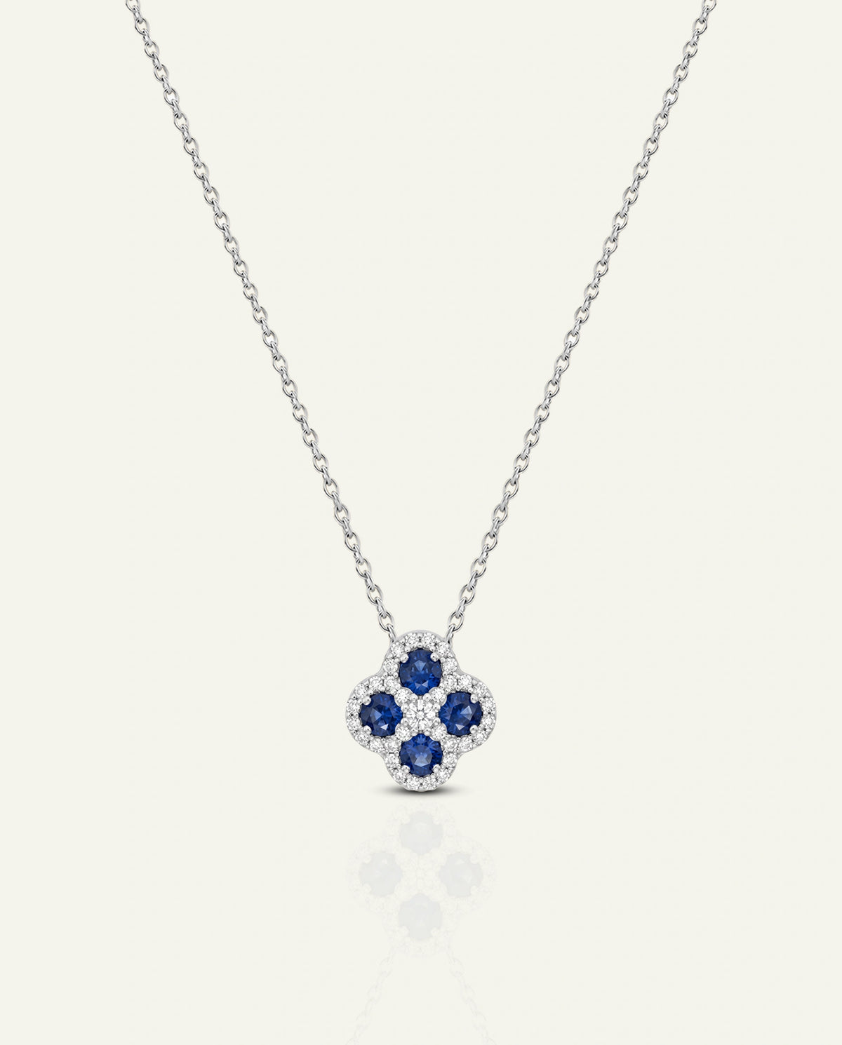 A silver-toned necklace with a clover-shaped pendant featuring four deep blue round-cut stones surrounded by small clear stones, displayed on a light cream background.