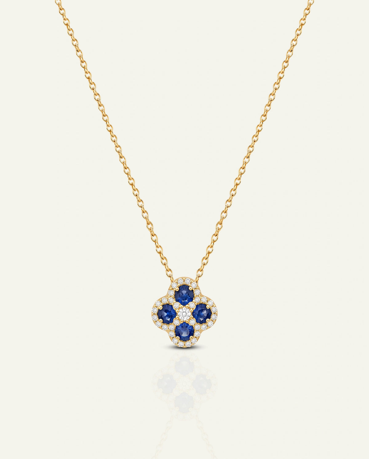 A yellow gold chain necklace featuring a four-petal clover motif pendant set with deep blue round-cut sapphires and surrounded by a halo of small clear pavÃ© diamonds, displayed on a light cream background.
