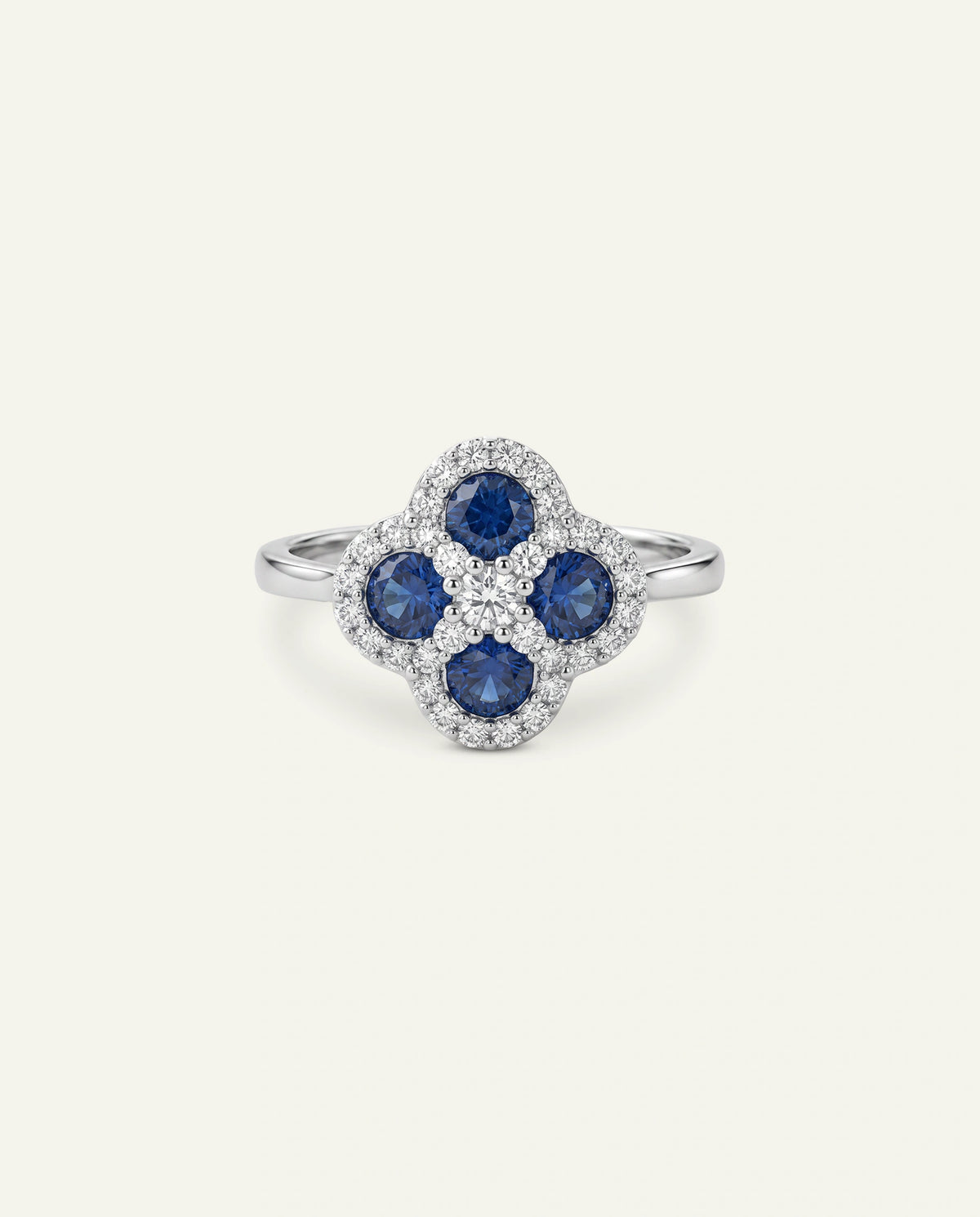 A silver-toned or white gold ring featuring a four-petal clover motif set with round blue sapphires, bordered by a halo of small clear pavÃ© diamonds, set against a light cream background.