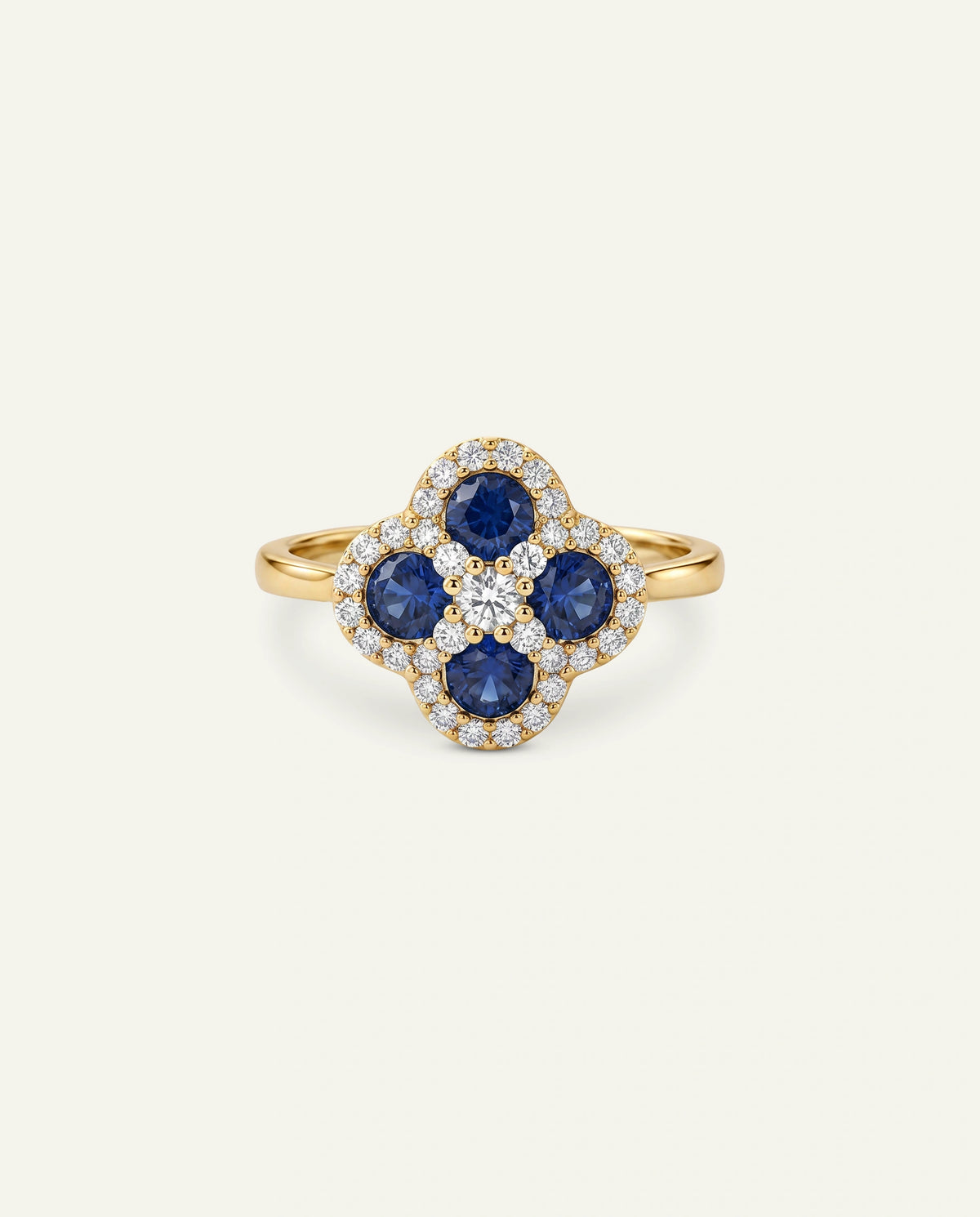 A gold ring featuring a clover-shaped floral cluster with four deep blue sapphires and a halo of small clear pavÃ© stones, set against a light cream background.