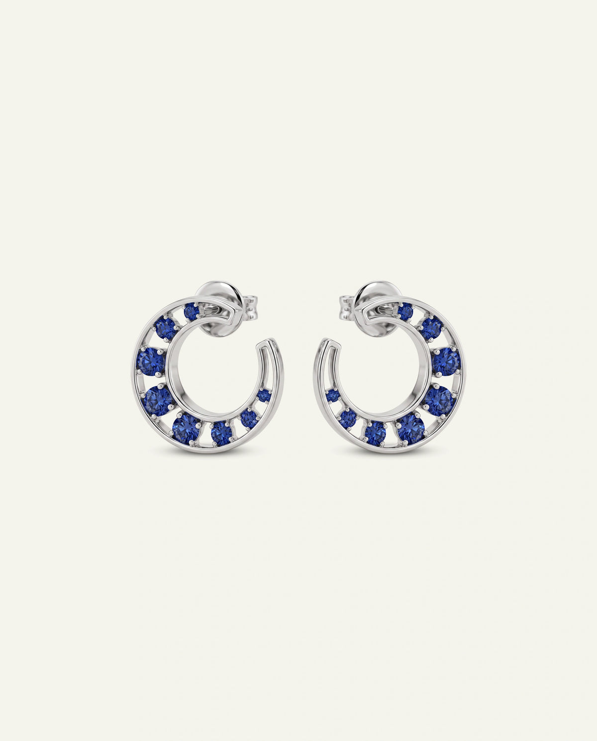A pair of silver-toned crescent-shaped hoop earrings adorned with small round blue gemstones, displayed on a light cream background.