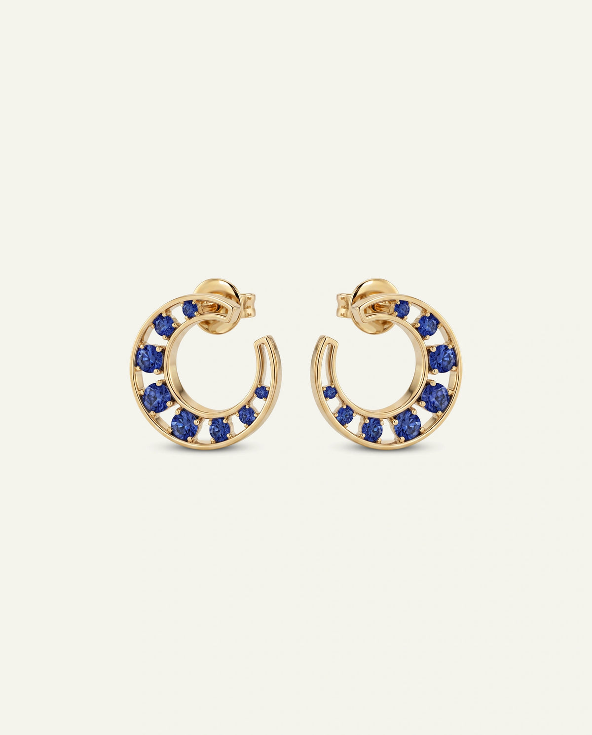 A pair of gold hoop earrings with small round blue gemstones set along the outer curve, shown on a plain light warm-gray background.