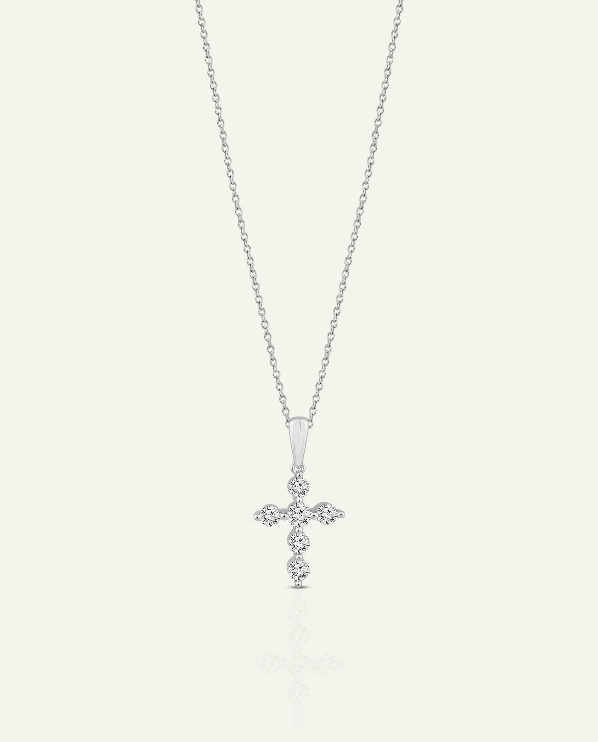 A delicate silver cross pendant necklace adorned with small sparkling stones, suspended from a fine silver chain against a light cream background.