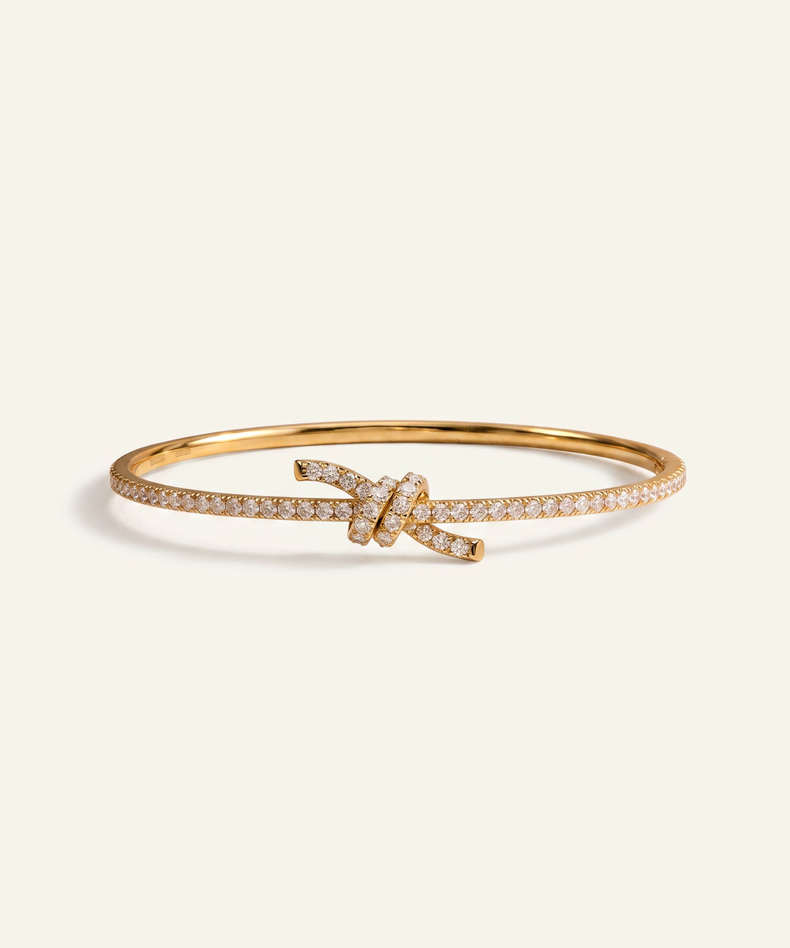 A gold bangle bracelet featuring a knotted center detail paved with small clear stones, set against a solid off-white background.