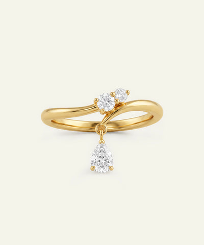 A yellow gold ring with a unique split-band design, featuring two round clear gemstones on one side and a pear-shaped clear gemstone hanging from a drop from the other side, set against a solid off-white background.