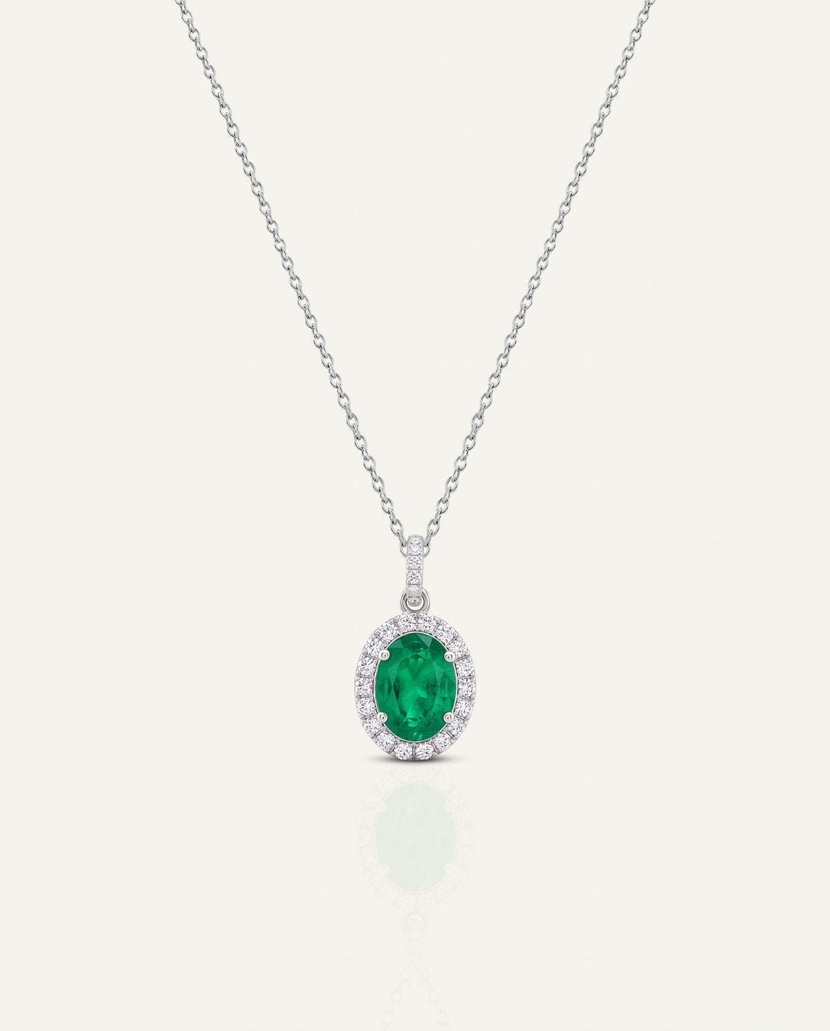 An oval emerald-cut green gemstone pendant necklace with a halo of small clear stones, suspended on a delicate silver-toned chain against a light cream background.