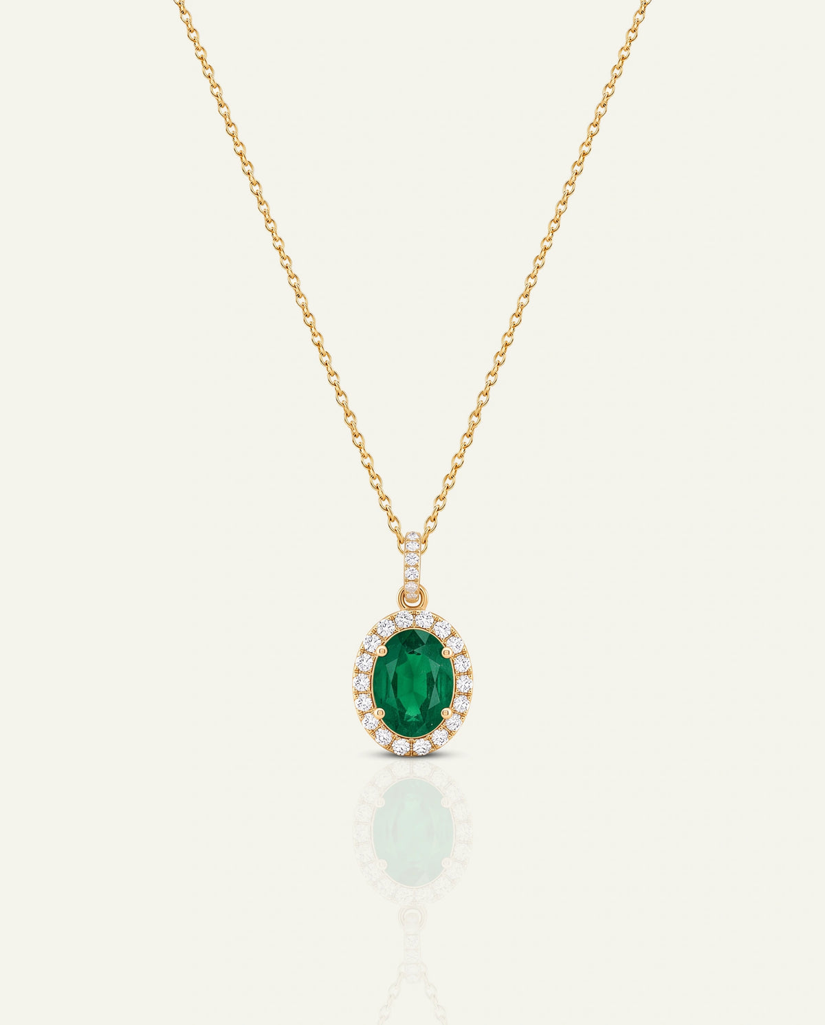 A delicate yellow gold chain necklace featuring an oval-cut green emerald pendant framed by a halo of small clear diamonds, set against a light cream background with a soft reflection.