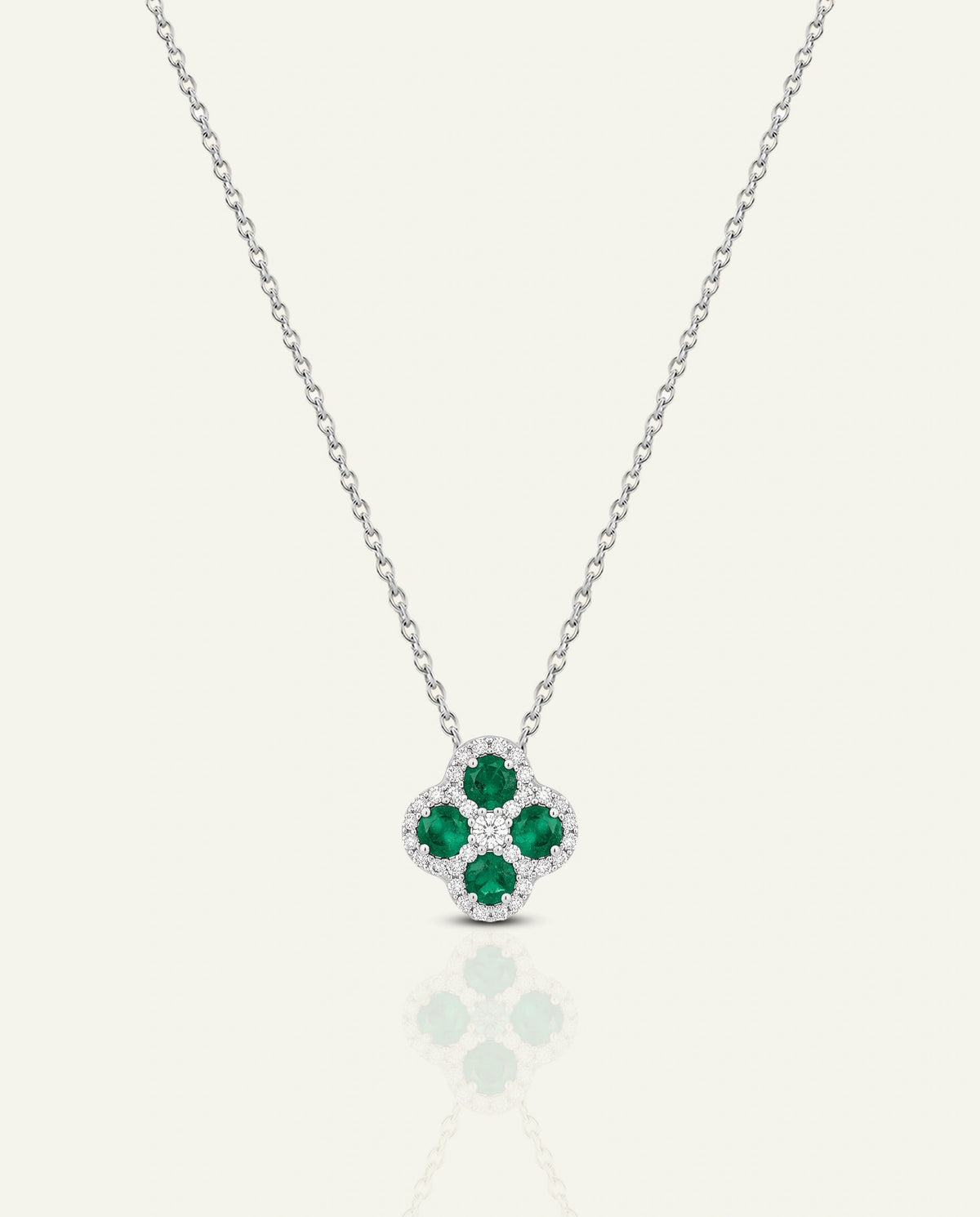 A silver-toned necklace with a four-petal flower pendant featuring three green emerald-cut gemstones and a halo of small clear stones, shown on a light cream background with a soft reflection below.