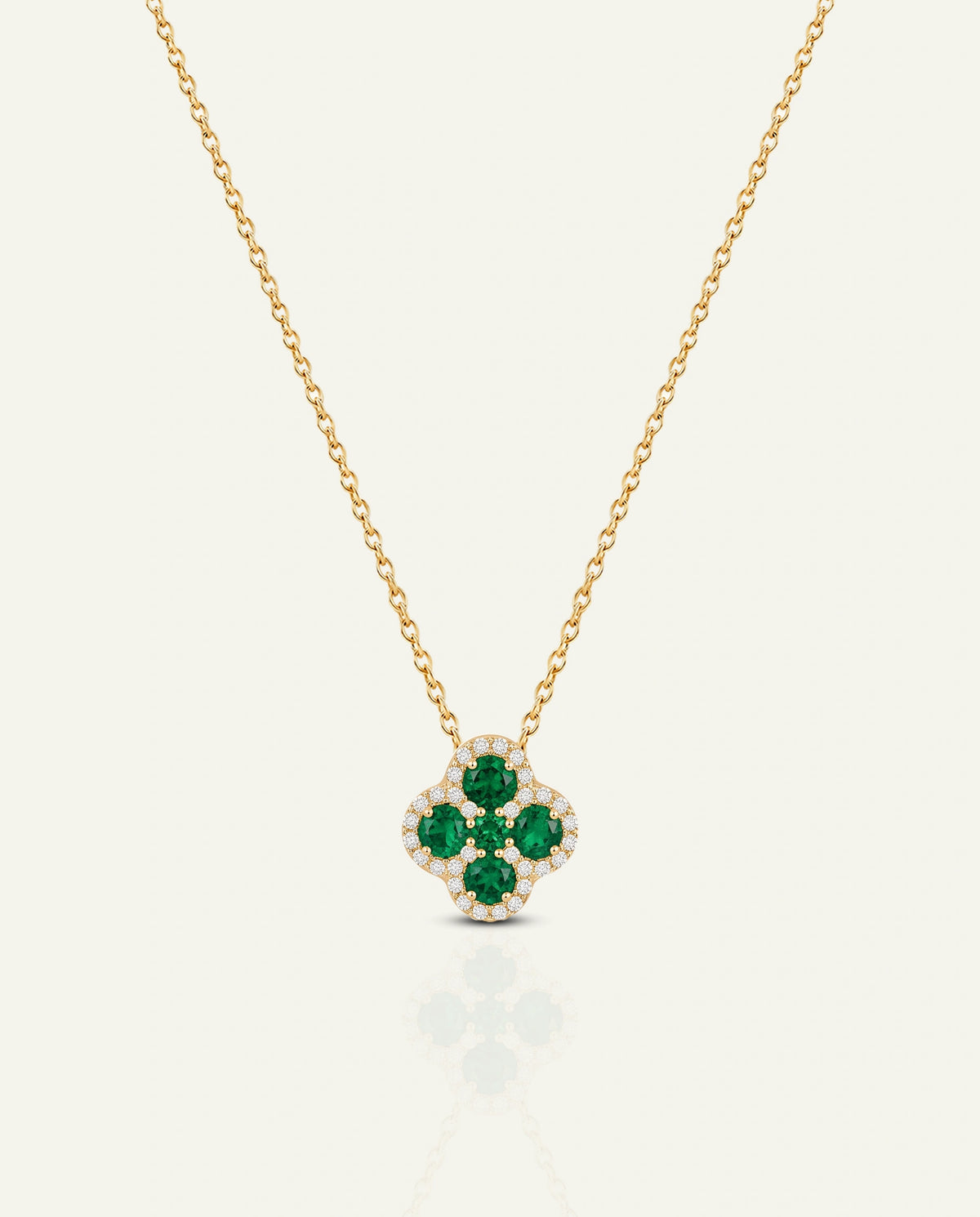 A yellow gold chain necklace featuring a four-petal clover motif pendant set with vibrant green emerald center stones and outlined by a halo of small clear diamonds, shown on an off-white background.